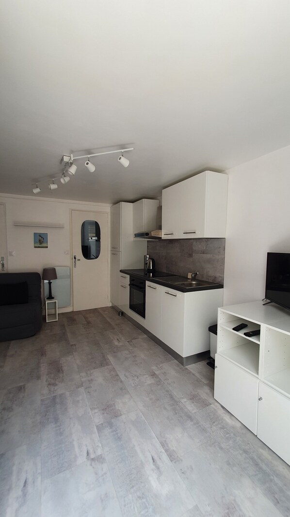 A modern kitchen with white cabinets, a black countertop, and a built-in oven. There is a small dining area with a table and chairs, and a TV on a stand.
