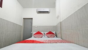 Ein schmales Hotelzimmer mit einem Bett, einer rot-weißen Tagesdecke, zwei rot-weißen Kissen und einer an der Wand montierten Klimaanlage.