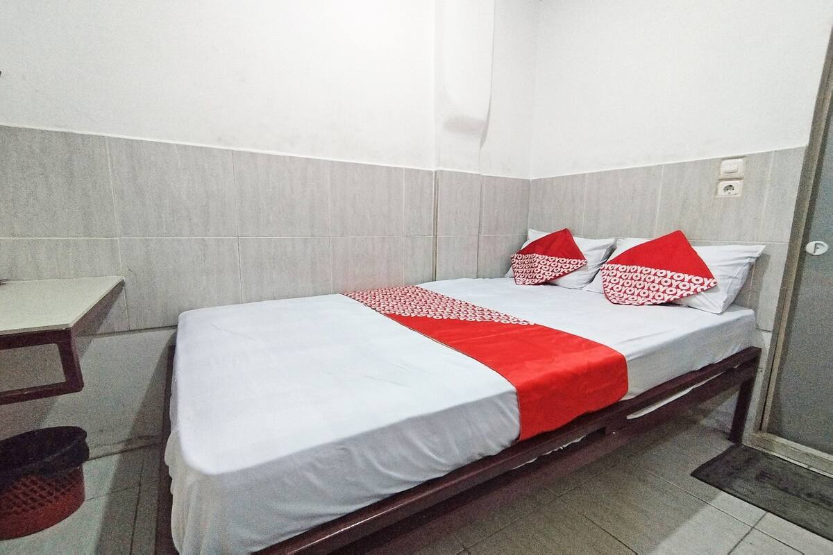 A single bed with a white and red bedspread, two pillows with red and white patterns, a wooden headboard, and a small bedside table.