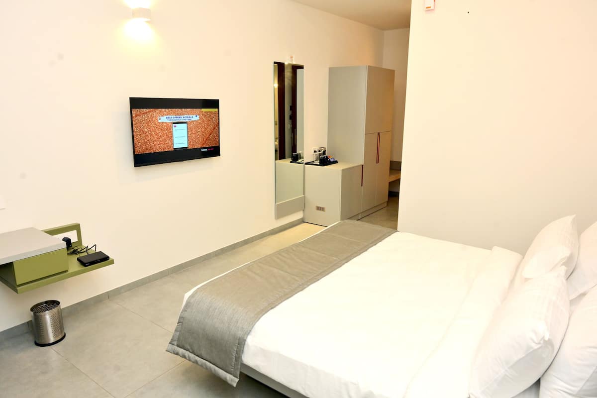 Une chambre d’hôtel comprenant un lit, un petit bureau avec une chaise, une poubelle, un miroir et un téléviseur fixé au mur qui affiche un menu.