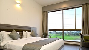A hotel room with a large bed, a desk, and a view of a green landscape through the window. - Clarks Inn Express, KRS Road, Mysore (Mysore)