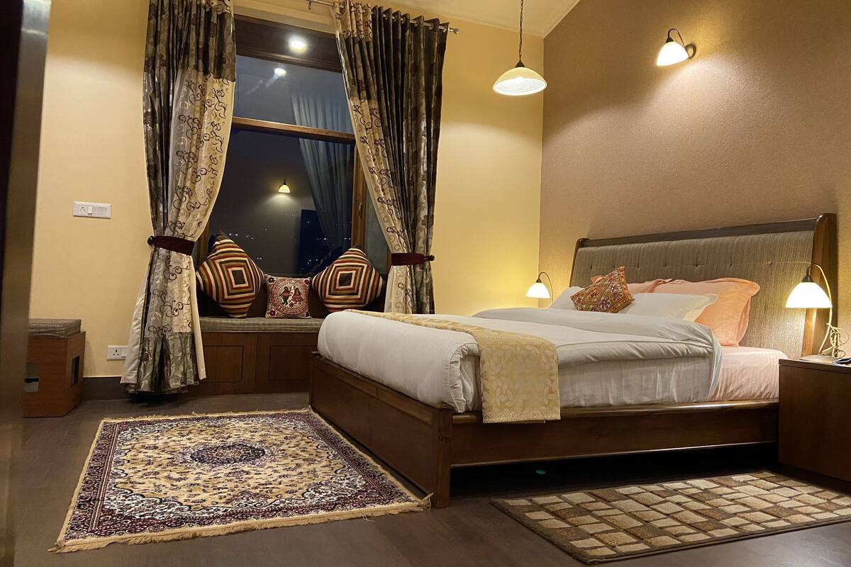 Ein Schlafzimmer mit einem großen Bett, zwei Nachttischlampen, einem Fenster mit Vorhängen, einem gemusterten Teppich und einem Nachttisch.