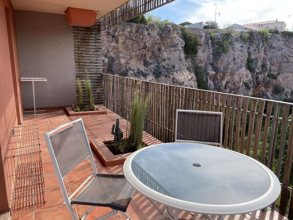 Een balkon met een ronde tafel en stoelen, een metalen grill en uitzicht op rotsachtige kliffen.