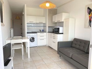 Um espaço de estar compacto com cozinha compacta, máquina de lavar, sofá cinzento e televisão.