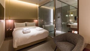 Et moderne hotelværelse med en stor seng, et glasafskærmet brusebad, et siddeområde og et badeværelse med spejl og vask.