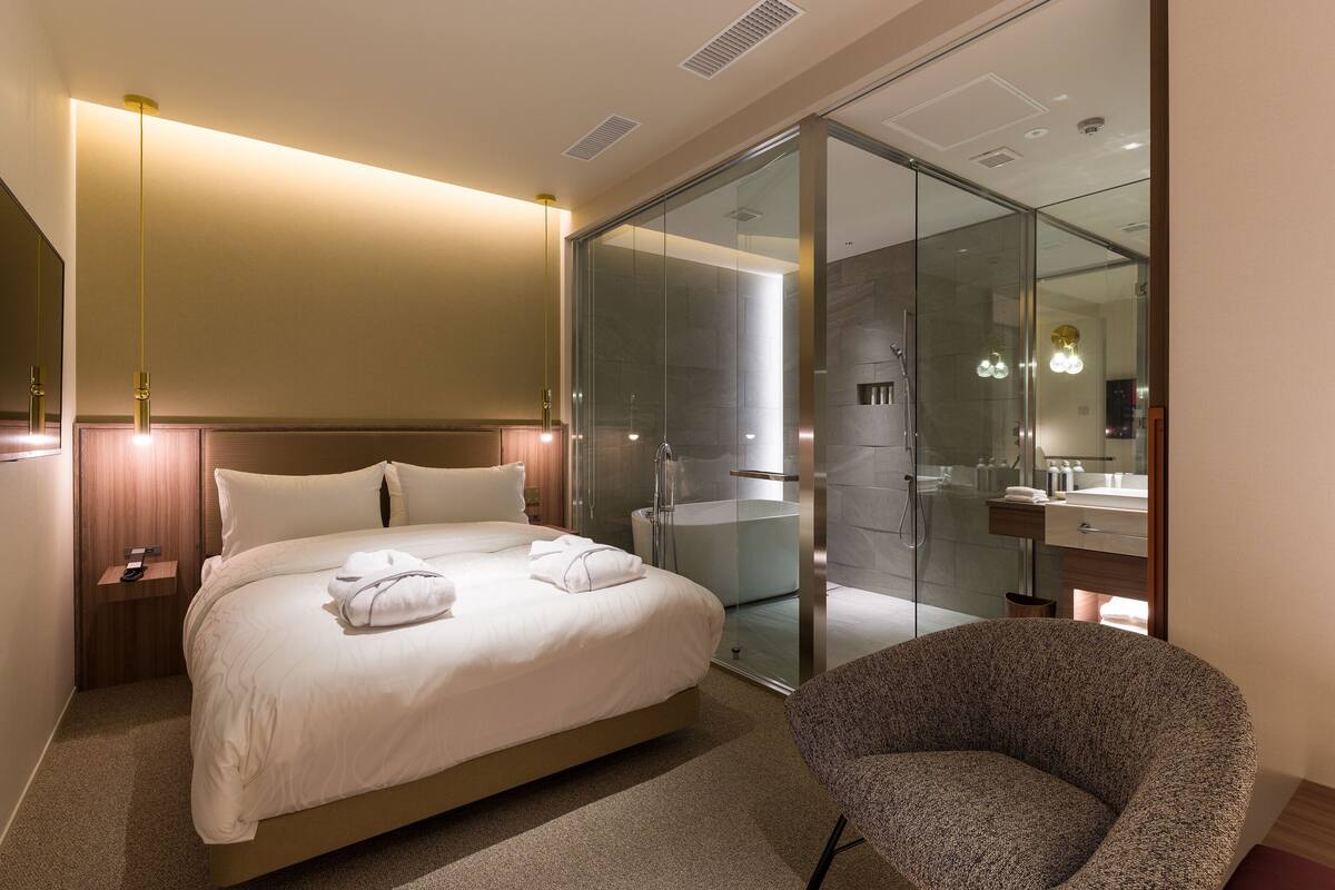 A modern hotel room with a large bed, a glass-enclosed shower, a sitting area, and a bathroom with a mirror and sink.