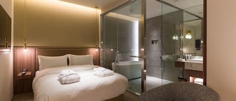 A modern hotel room with a large bed, a glass-enclosed shower, a sitting area, and a bathroom with a mirror and sink.