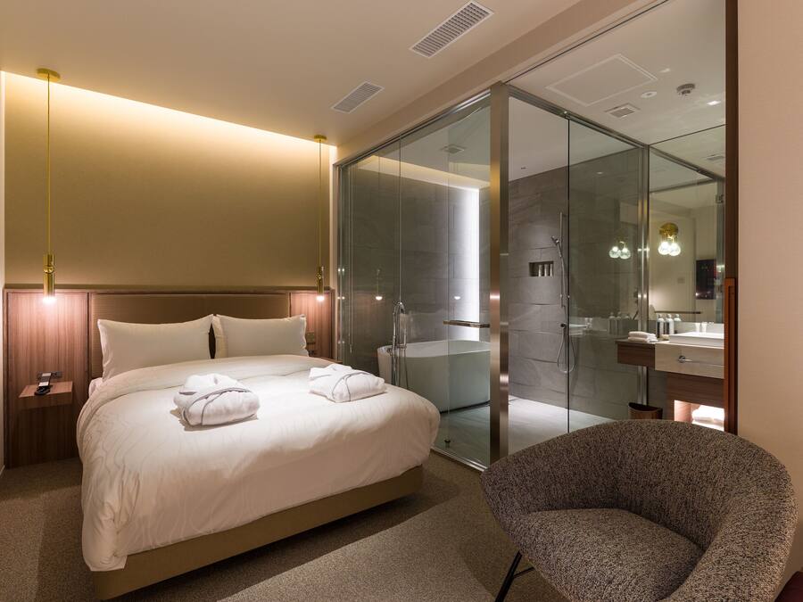 A modern hotel room with a large bed, a glass-enclosed shower, a sitting area, and a bathroom with a mirror and sink.