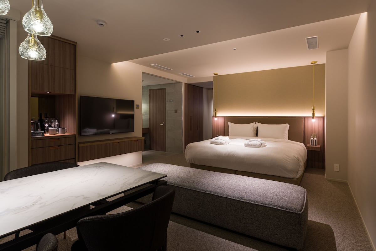 A modern hotel room with a large bed, a desk with a chair, a television, and a minibar.