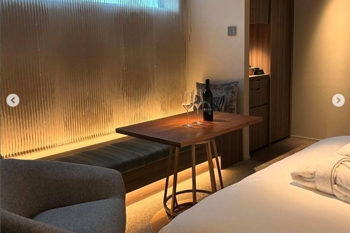 A hotel room with a bed, a wooden table with a bottle and glasses, a bench, and a window with curtains.