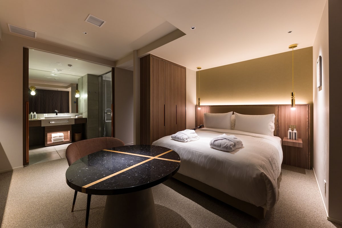 A modern hotel room with a bed, a round table, a bathroom with a mirror, and a nightstand with toiletries.