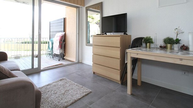A modern living room with a sofa, a wooden cabinet, a TV on a stand, a desk with plants, and a large sliding glass door leading to an outdoor area. - Bright Studio with Terrace, Near Shops and Thermal Baths (Balaruc-les-Bains)