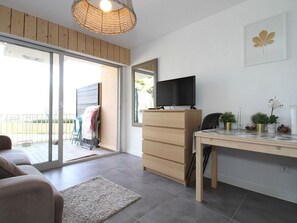 A modern living room with a sofa, a wooden cabinet, a TV on a stand, a desk with plants, and a large sliding glass door leading to an outdoor area. - Bright Studio with Terrace, Near Shops and Thermal Baths (Balaruc-les-Bains)