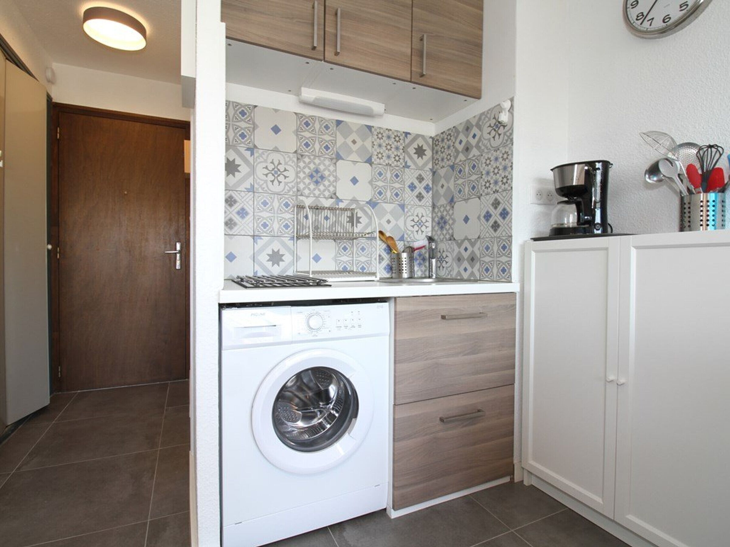 Une cuisine compacte dotée d’un lave-linge encastré, de meubles en bois et d’un pare-feu en carrelage.