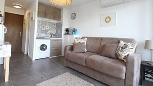 A modern living room with a brown sofa, a woven pendant light, and a kitchen in the background. - Bright Studio with Terrace, Near Shops and Thermal Baths (Balaruc-les-Bains)