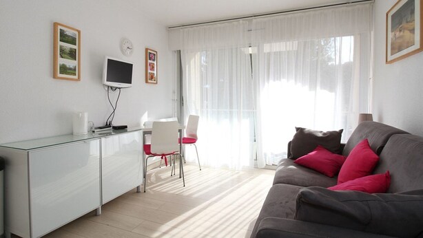 A modern living room with a grey sofa, a glass desk, a white chair, and a TV on the wall. - Modern Studio with Terrace, AC, and Parking (Balaruc-les-Bains)