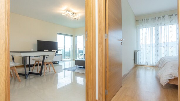 A modern hotel room with a wooden door, a bed, a desk with chairs, a television, and a large window with curtains. - Buarcos Sunset Apartment by Rent4All (Figueira da Foz)