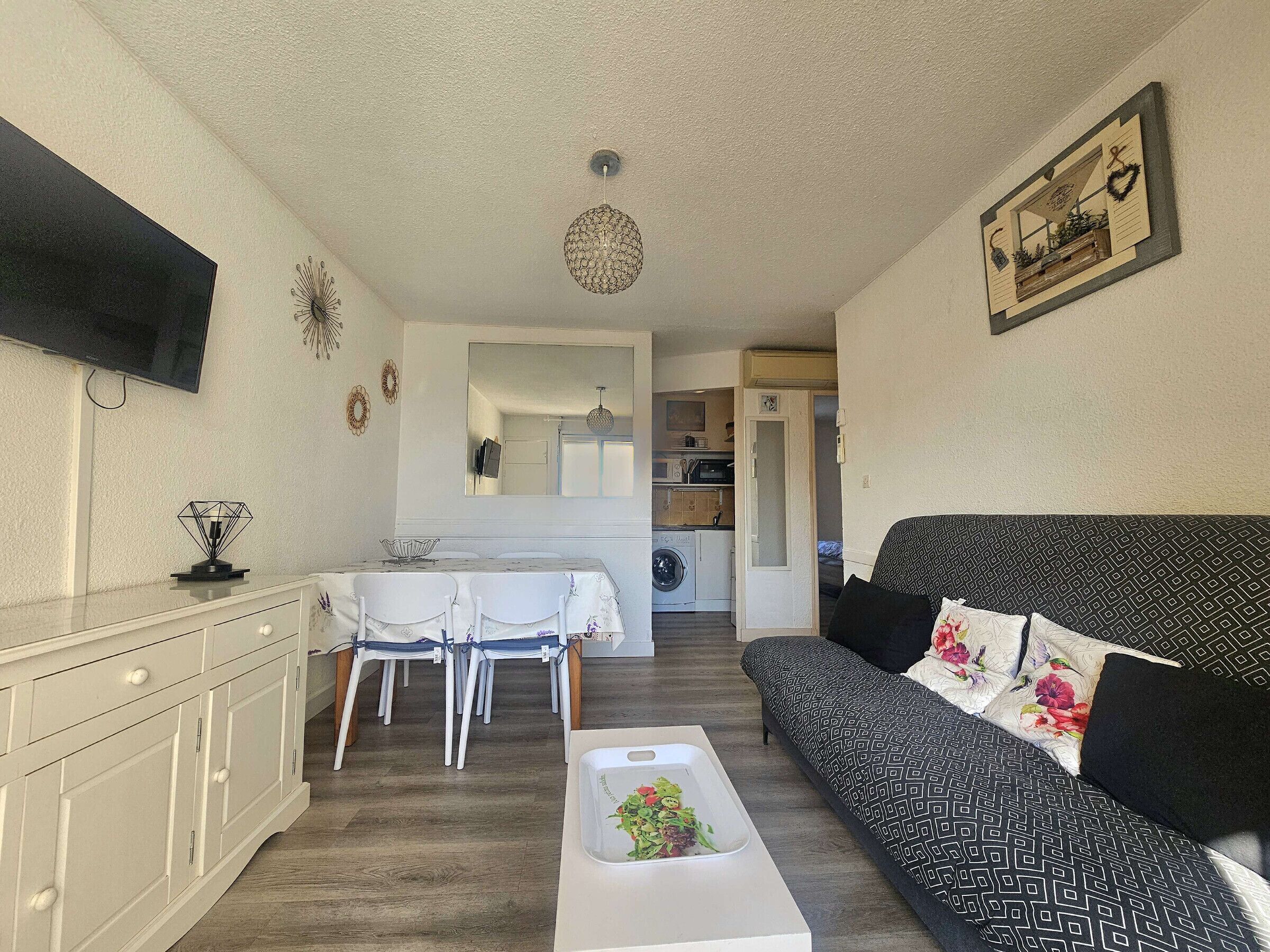Een woonkamer met een bank, een salontafel met een plant, een eethoek met stoelen en een tafel, en een keuken op de achtergrond.