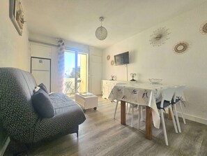 A living room with a patterned armchair, a dining table with chairs, a TV mounted on the wall, and a balcony with a sliding door. - Beautiful 1BR with Parking, AC and Wifi - Balaruc-les-Bains (Balaruc-les-Bains)