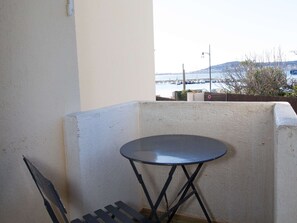 Un balcon avec une petite table ronde et une chaise, donnant sur une étendue d’eau où l’on aperçoit des bateaux et, au loin, un pont.