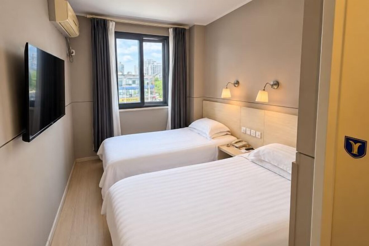 A hotel room with two beds, a TV, a window with curtains, and a view of the city.