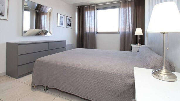 Un dormitorio moderno con una cama grande, un aparador, una mesita de noche con lámpara, una ventana con cortinas y un espejo en la pared.