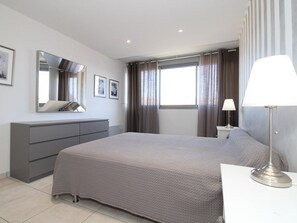 Un dormitorio moderno con una cama grande, un aparador, una mesita de noche con lámpara, una ventana con cortinas y un espejo en la pared.
