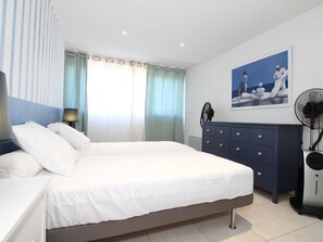 A bedroom with a large bed, a dresser, a fan, and a painting on the wall.
