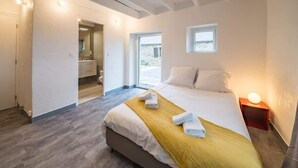 Un dormitorio con cama, una mesita de noche con lámpara, una ventana y un baño visible a través de una puerta abierta.