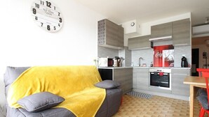 A modern living room with a sofa, a yellow throw blanket, and a kitchen area with a microwave, sink, and a red Coca-Cola sign. - Renovated T2 with Terrace Near Thau Lagoon, 800m from Thermes, Parking, Wifi, A/C (Balaruc-les-Bains)