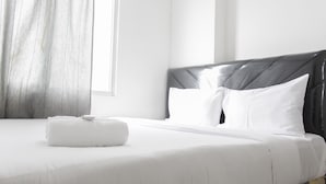 A neatly made bed with white linens and pillows, a grey headboard, and a window with a curtain.