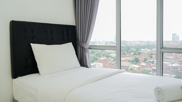 Ein ordentlich bezogenes Bett mit schwarzem Kopfteil und weißer Bettwäsche, in einem Zimmer mit großen Fenstern und Blick auf die Stadt.