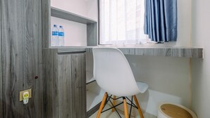 A room with a wooden door, a white chair, a shelf with water bottles, a window with blue curtains, and a wooden floor. - Great Choice And Comfortable Studio Transpark Cibubur Apartment (Depok)