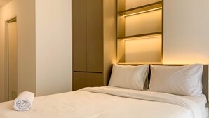 Una cama bien hecha con sábanas blancas y una toalla enrollada, un armario de madera e iluminación empotrada en la pared.