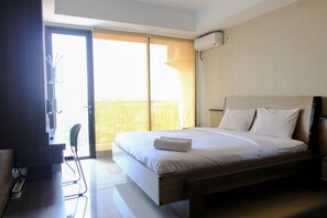 A hotel room with a bed, a desk with a chair, a window, and an air conditioning unit. - Homey Studio Room At 6Th Floor Beverly Dago Apartment (Bandung)