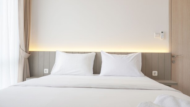 Cama bem arrumada com lençóis e almofadas brancas, mesa de cabeceira com abajur e termostato fixado na parede.