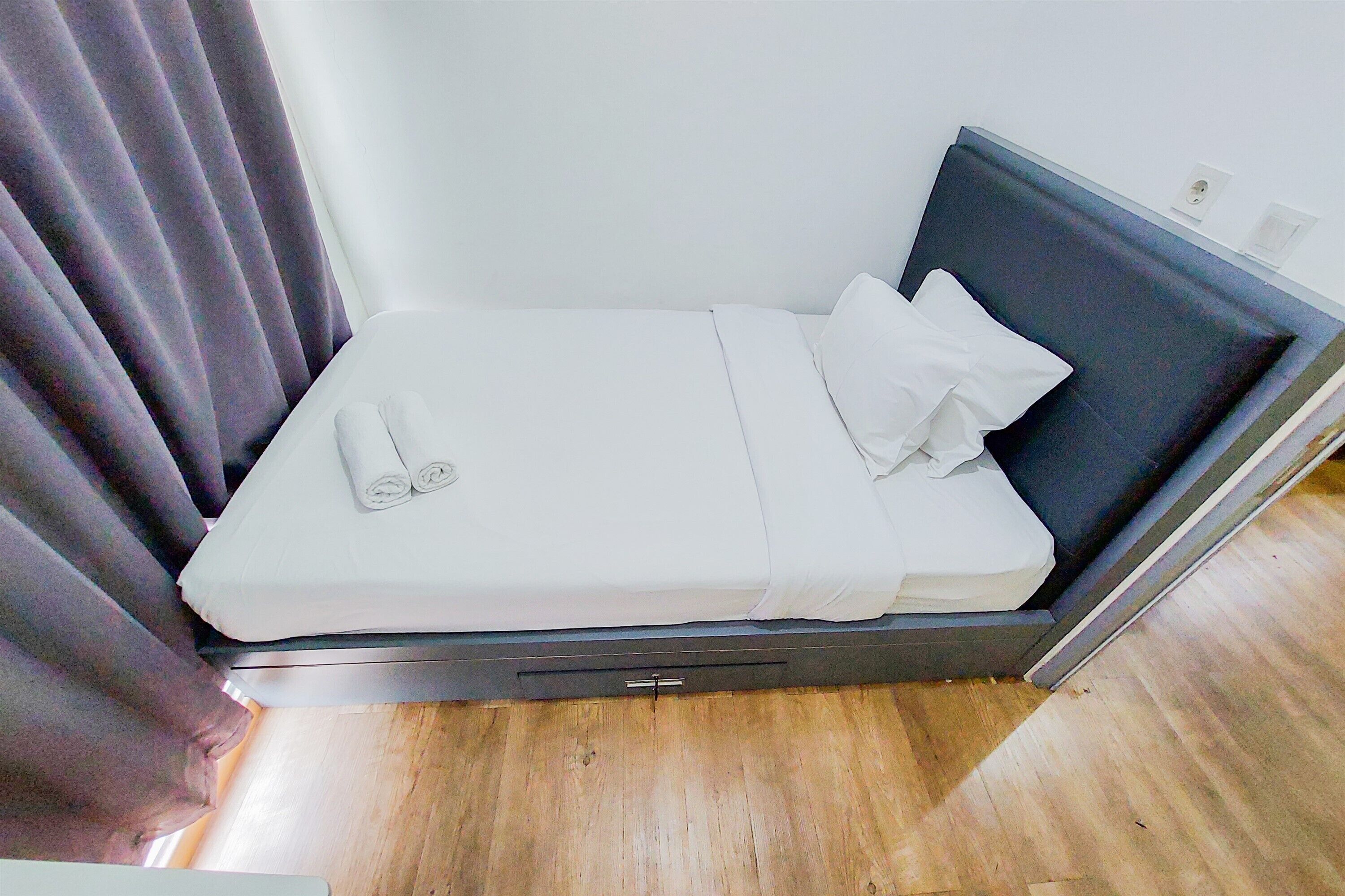 Una cama pequeña y ordenada con sábanas blancas y cabecera oscura en una habitación con suelo de madera y cortina oscura.