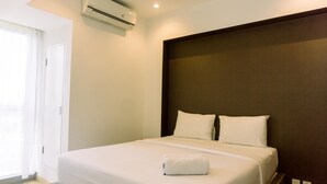 A hotel room with a bed, white bedding, a wooden headboard, an air conditioning unit, and a window with curtains. - Nice And Fancy 1Br At Branz Bsd City Apartment (Pagedangan)