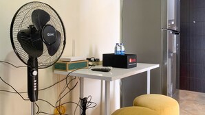 Una stanza piccola con un ventilatore portatile, un tavolo con una scatola e un telecomando, una bottiglia d'acqua e un poggiapiedi giallo.