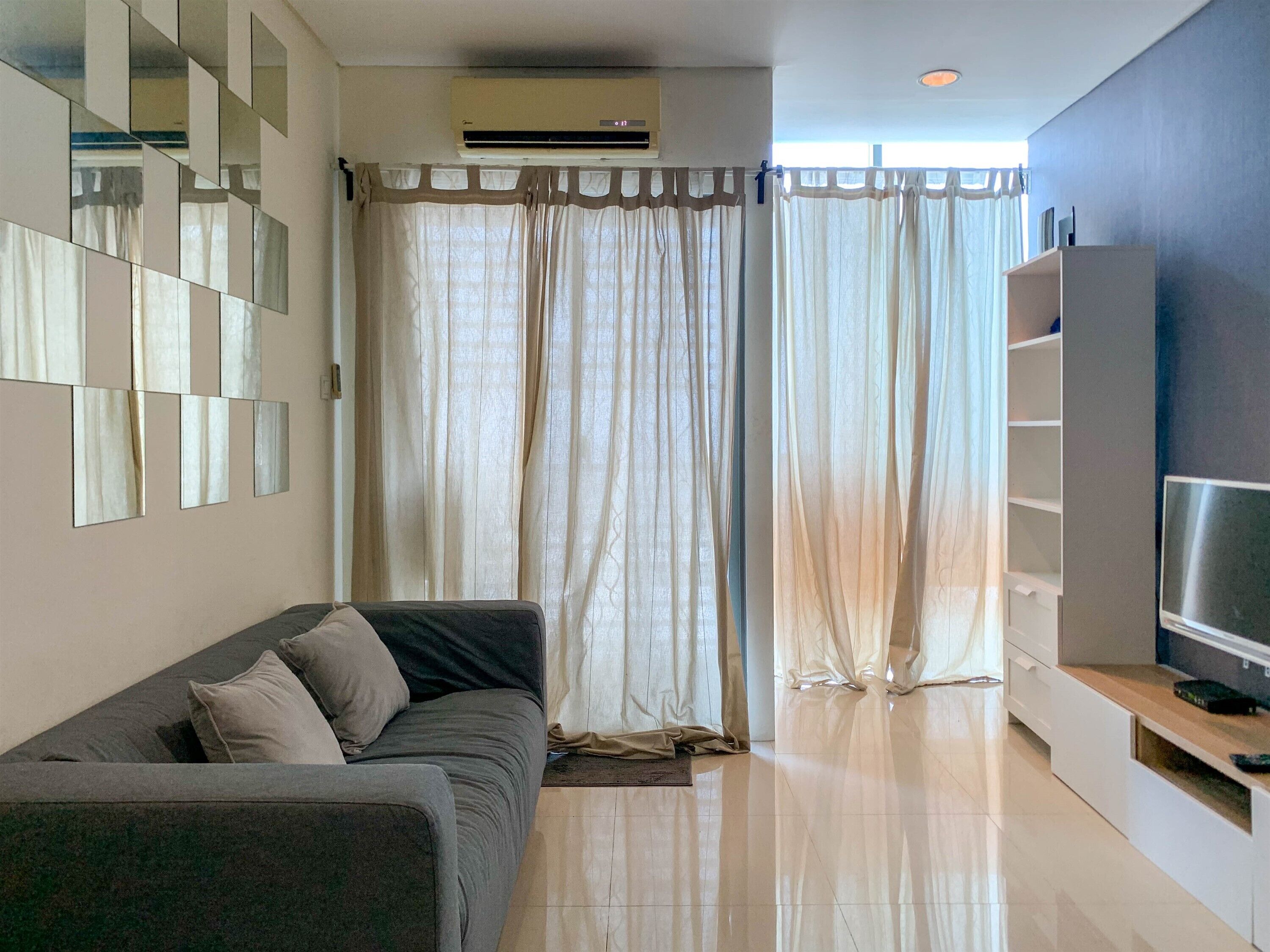 A modern living room with a grey sofa, a TV on a wooden stand, and a large window with sheer curtains.