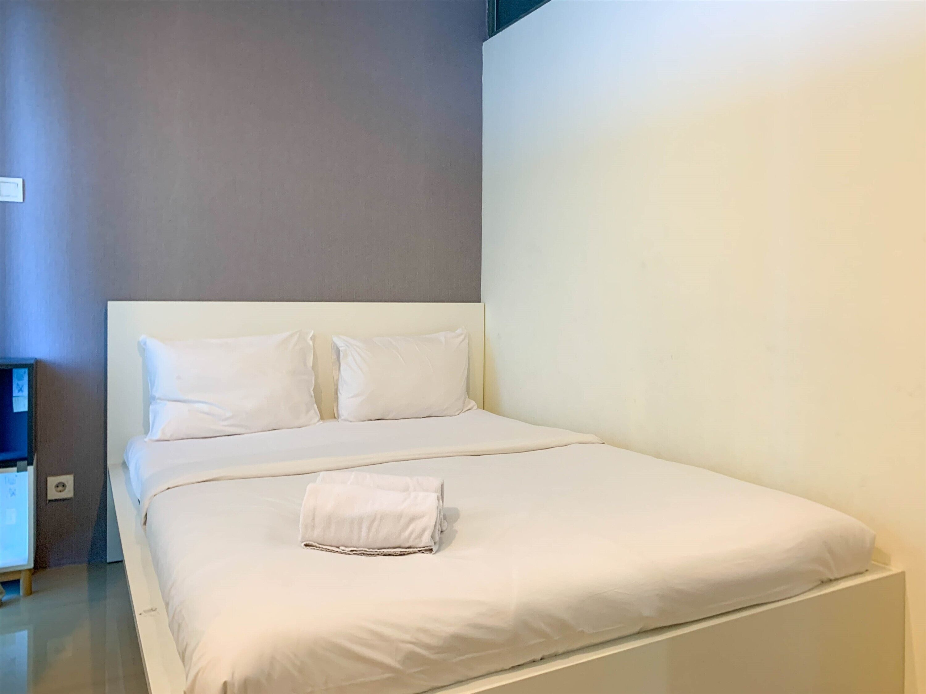 Una cama bien hecha con sábanas y almohadas blancas, una toalla doblada sobre la cama y una mesita de noche con una lámpara.