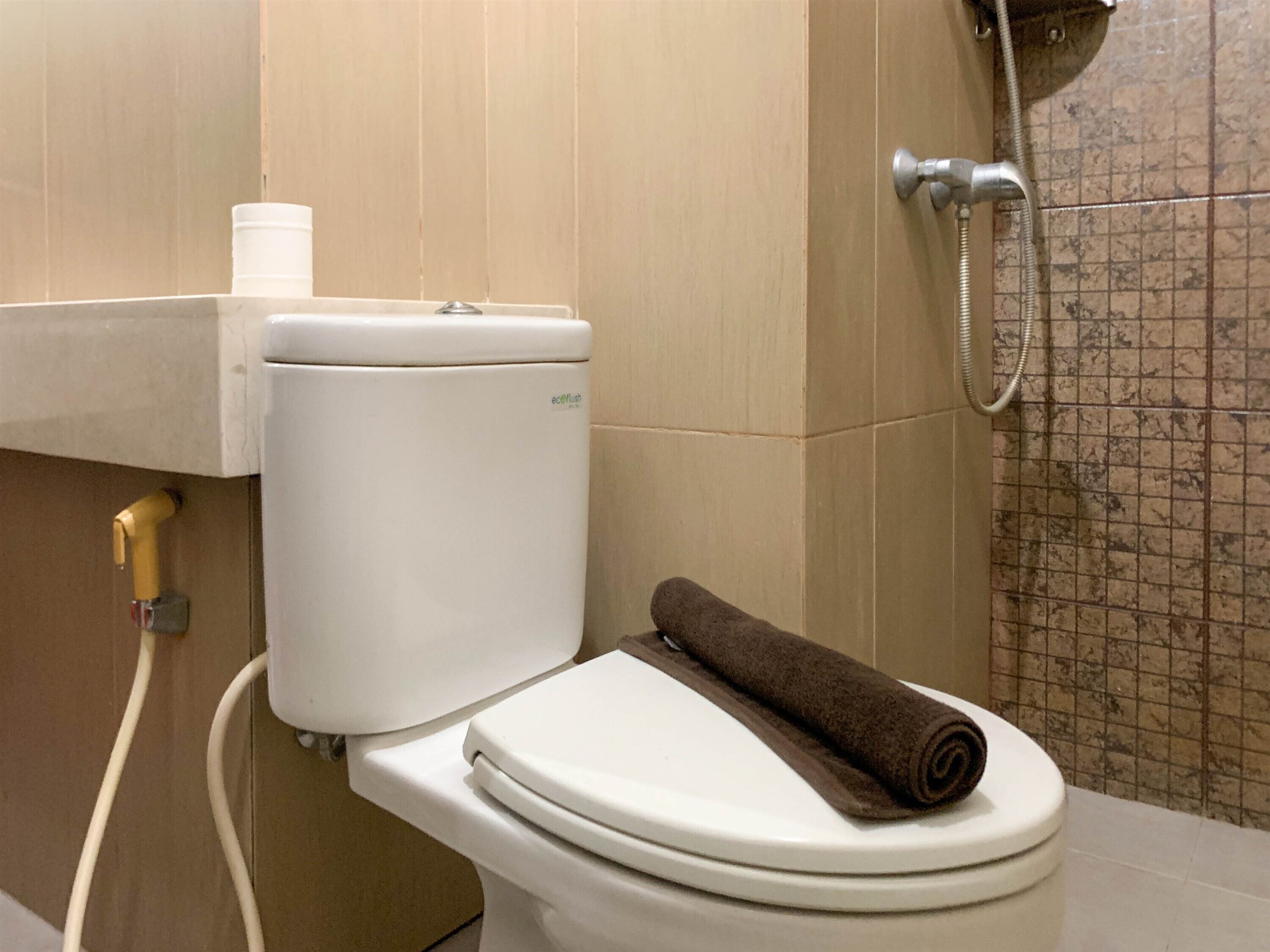 Inodoro blanco con una toalla marrón encima, un dispensador de papel higiénico y una zona de ducha con pared de azulejos.