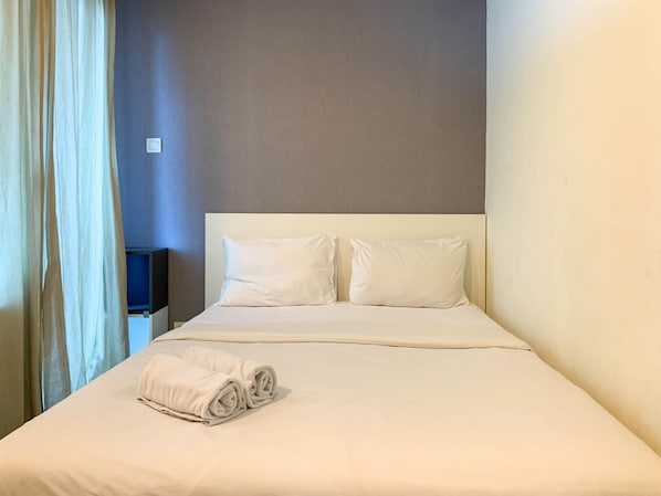 A neatly made bed with white linens and pillows, a folded towel on the bed, and a plain wall with a light switch.