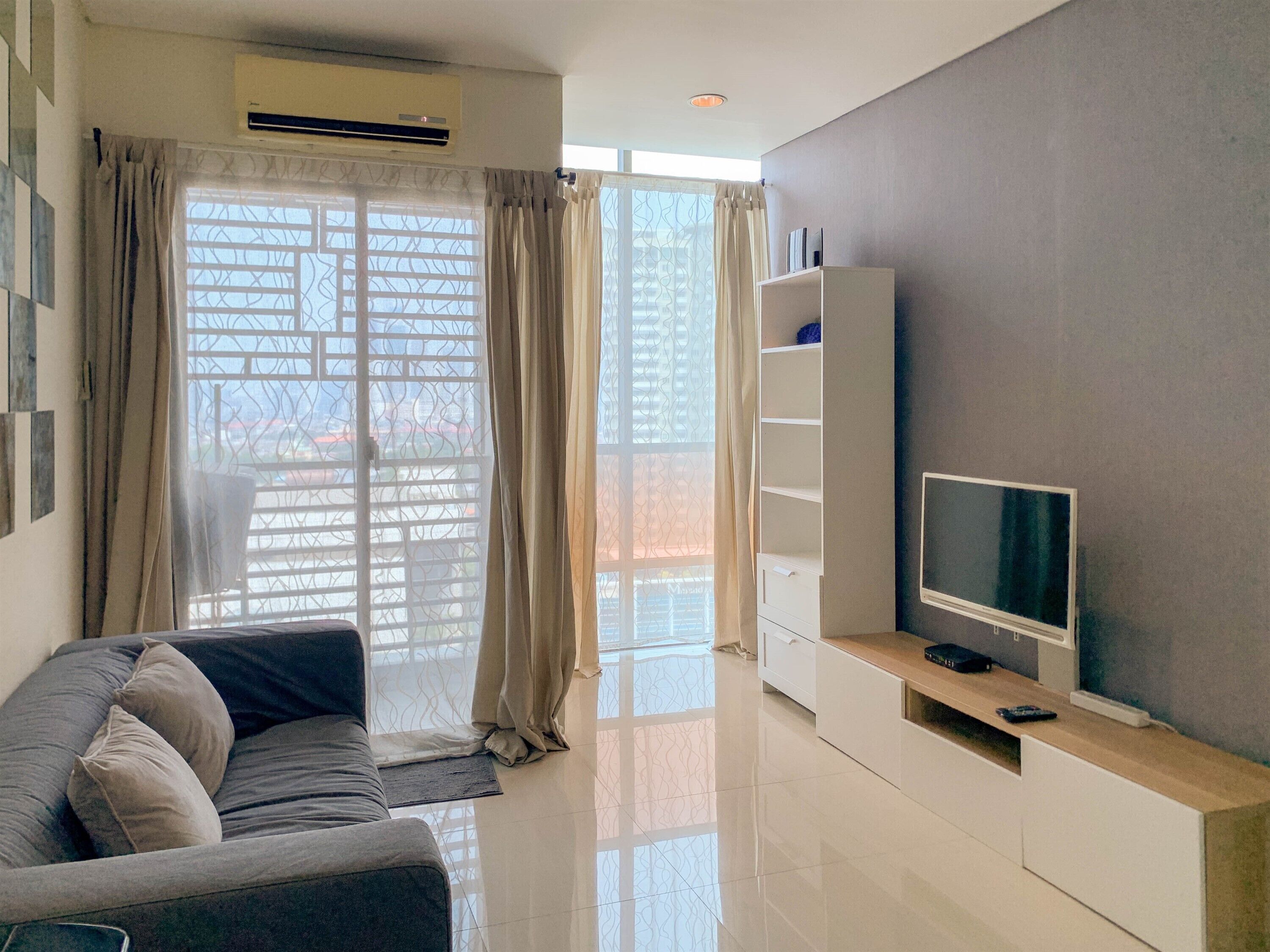 Una sala moderna con un sofá gris, un televisor sobre un mueble de madera y un ventanal con cortinas.