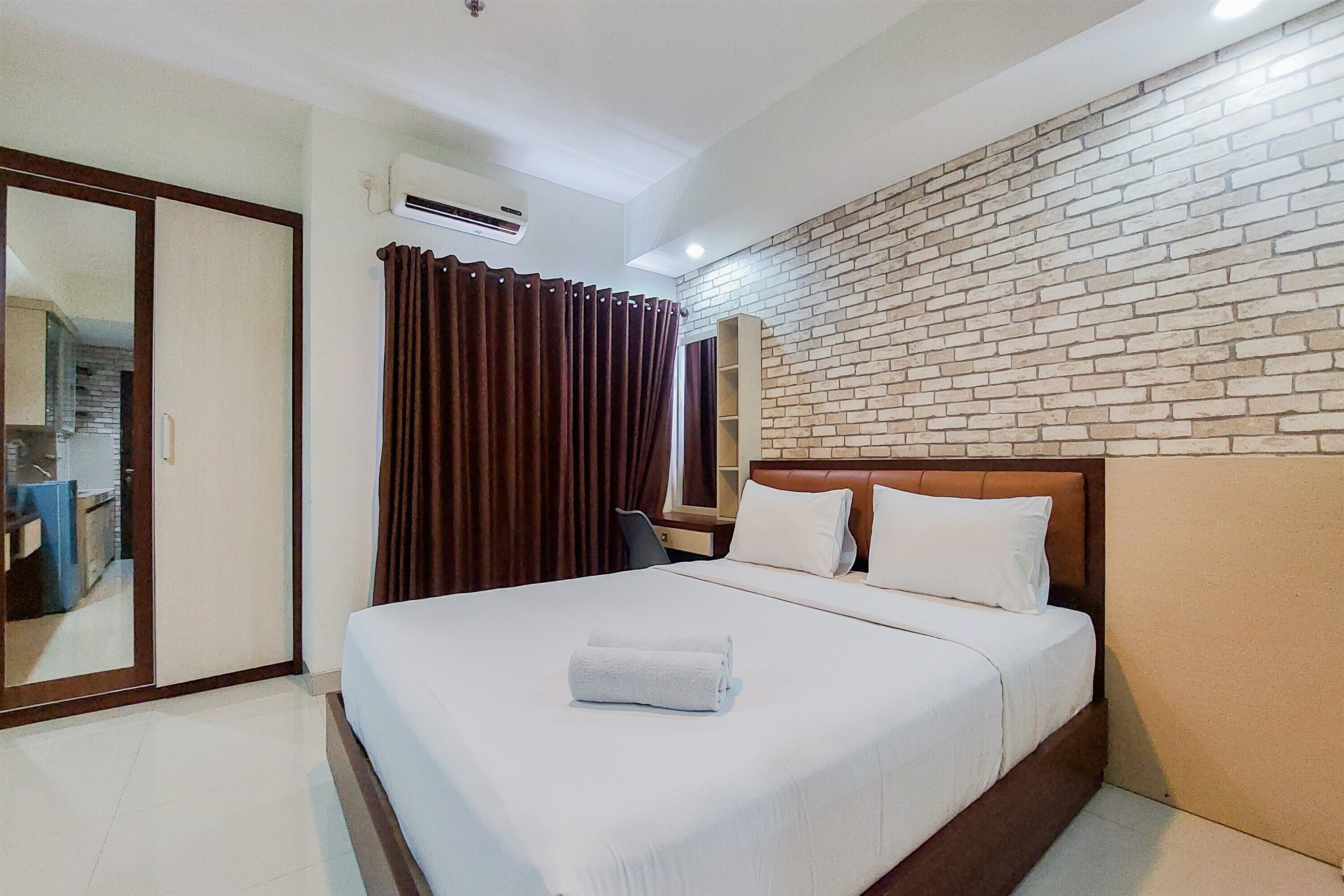 Quarto de hotel com cama grande, cabeceira de madeira, parede com padrão de tijolos, espelho e janela com cortinas.