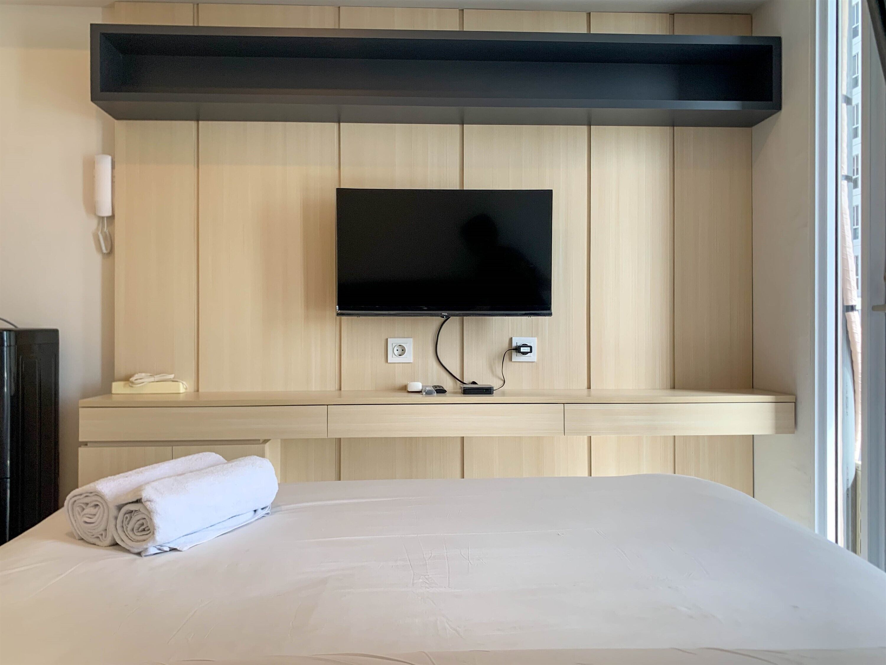 Een hotelkamer met een bed, een flatscreen televisie bevestigd aan een houten wand, een nachtkastje met een telefoon en een klein doosje, en een raam met gordijnen.