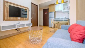 Un salon moderne avec un téléviseur, une table basse ronde, un canapé agrémenté d’un coussin rouge et un coin cuisine doté de meubles en bois et d’un réfrigérateur.
