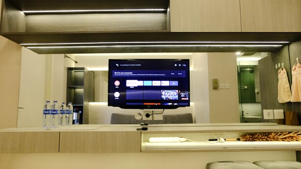 A modern hotel room with a large flat-screen TV mounted on the wall, a built-in desk with a keyboard and mouse, and a comfortable seating area with a cushioned bench. - Homey 1Br At Apartment Parahyangan Residence (Bandung)