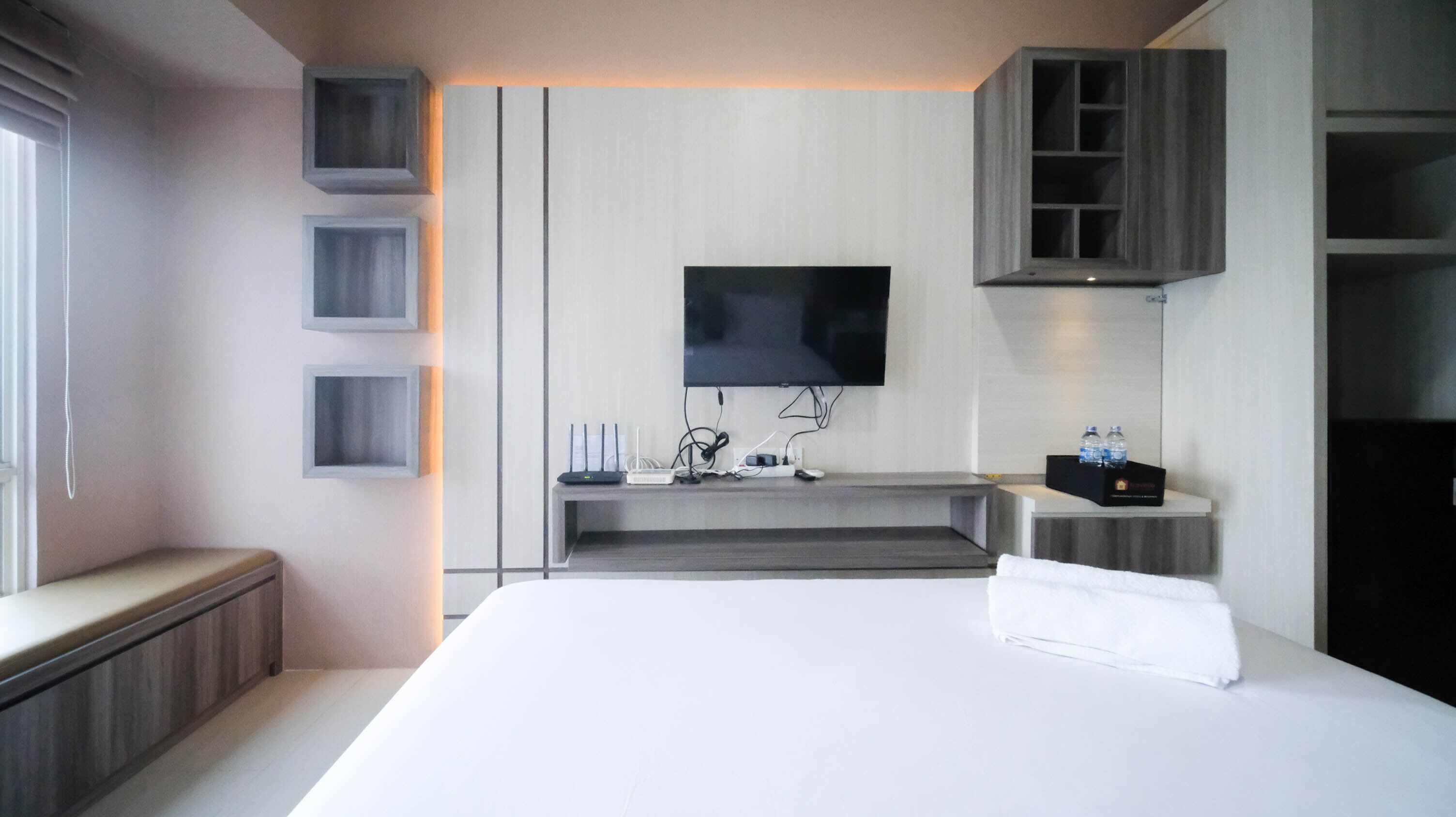 Una habitación de hotel moderna con una cama grande, un televisor de pantalla plana en la pared, un escritorio pequeño con una computadora y una botella de agua, y armarios empotrados.