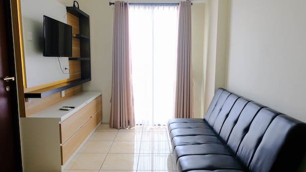 A modern living room with a black leather sofa, a flat-screen TV mounted on a wooden shelf, a sliding door with curtains, and a tiled floor. - Cozy And Homey 2Br Tamansari Panoramic Apartment (Bandung)